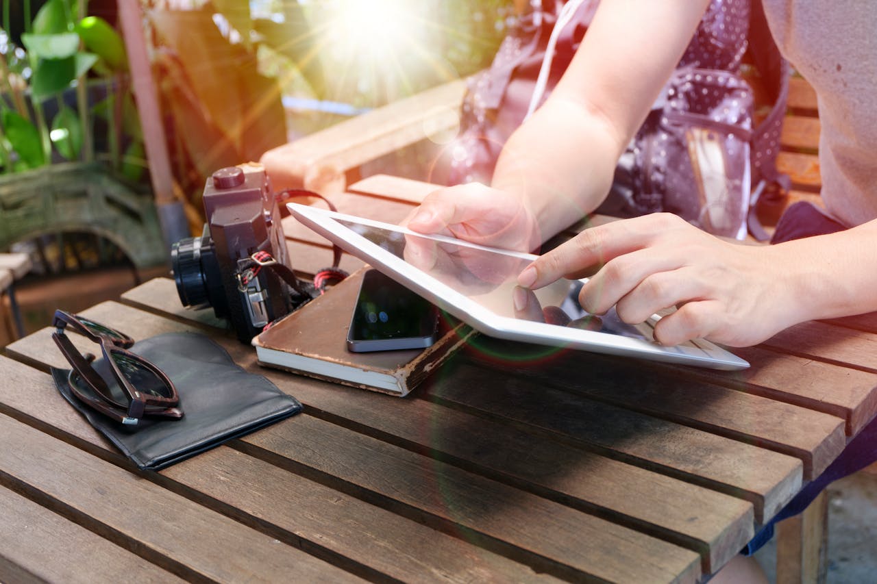 creative-02 Person using a tablet outdoors alongside a camera, phone, sunglasses, and notebook on a wooden table.